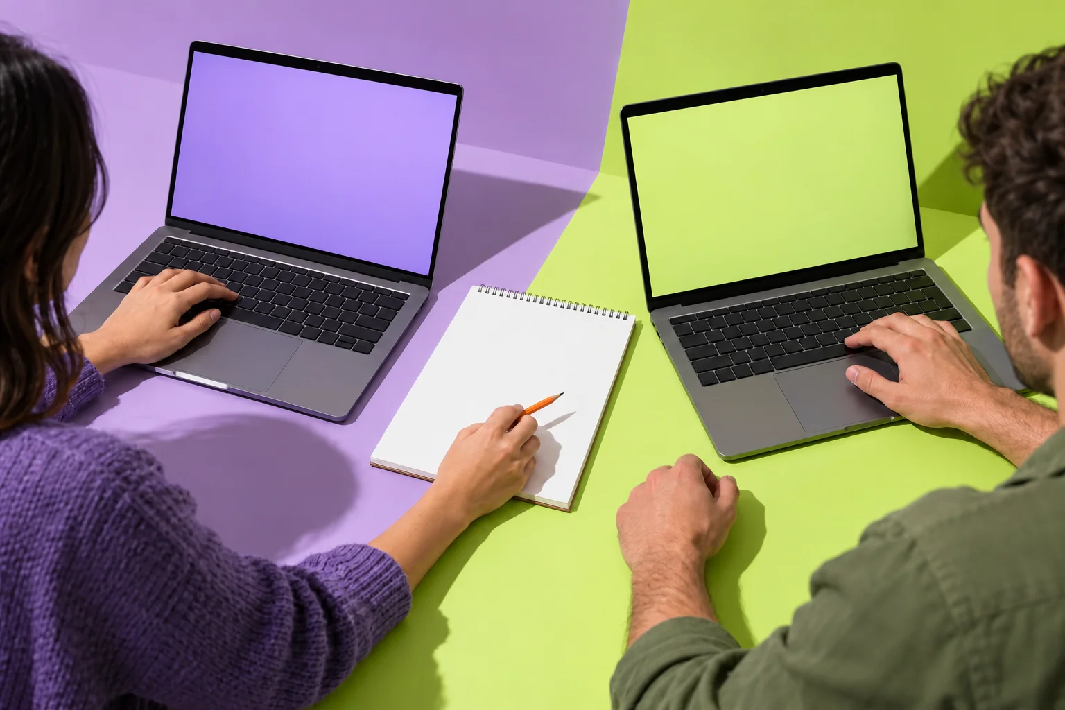 Studio photograph of two open laptops side by side with a shared notepad and an orange pencil.